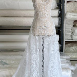 Peut inclure: Un tissu en dentelle blanche recouvrant un mannequin de couture. Le tissu pr&eacute;sente un motif floral complexe avec des bords festonn&eacute;s, mettant en valeur une texture d&eacute;licate et transparente qui s'&eacute;tend de l'encolure jusqu'&agrave; l'ourlet au sol.