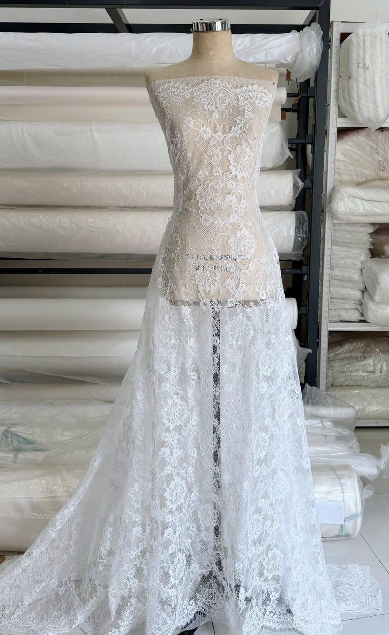 Peut inclure: Un tissu en dentelle blanche recouvrant un mannequin de couture. Le tissu pr&eacute;sente un motif floral complexe avec des bords festonn&eacute;s, mettant en valeur une texture d&eacute;licate et transparente qui s'&eacute;tend de l'encolure jusqu'&agrave; l'ourlet au sol.