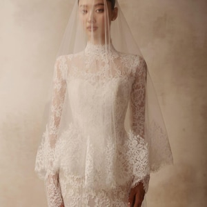Peut inclure: Une robe de mari&eacute;e en dentelle blanche avec un col montant, des manches longues et un d&eacute;tail de basque &agrave; la taille. Le v&ecirc;tement est orn&eacute; de motifs floraux complexes sur tout le corsage et les manches, accompagn&eacute; d'un voile de mari&eacute;e transparent.