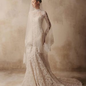 Peut inclure: Une robe de mari&eacute;e en dentelle blanche longue jusqu'au sol avec un col montant, des manches longues et un corsage ajust&eacute; qui s'&eacute;vase en tra&icirc;ne. La robe est orn&eacute;e de motifs floraux complexes et accompagn&eacute;e d'un voile en tulle transparent.