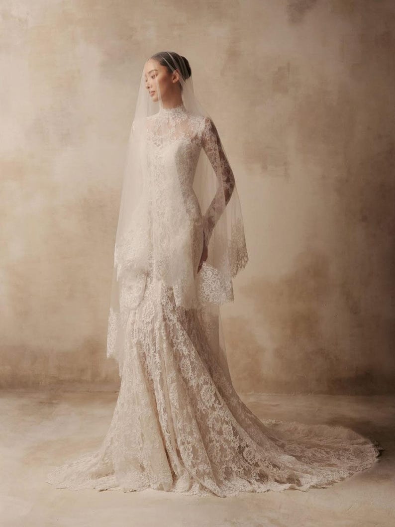 Peut inclure: Une robe de mari&eacute;e en dentelle blanche longue jusqu'au sol avec un col montant, des manches longues et un corsage ajust&eacute; qui s'&eacute;vase en tra&icirc;ne. La robe est orn&eacute;e de motifs floraux complexes et accompagn&eacute;e d'un voile en tulle transparent.