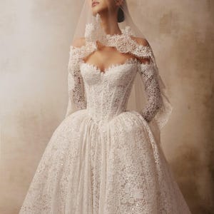Peut inclure: Une robe en dentelle blanche dot&eacute;e d'un corsage structur&eacute;, de manches longues et d'une encolure d&eacute;gag&eacute;e aux bords festonn&eacute;s. La jupe volumineuse est confectionn&eacute;e en dentelle florale complexe avec de fines paillettes et un voile transparent.