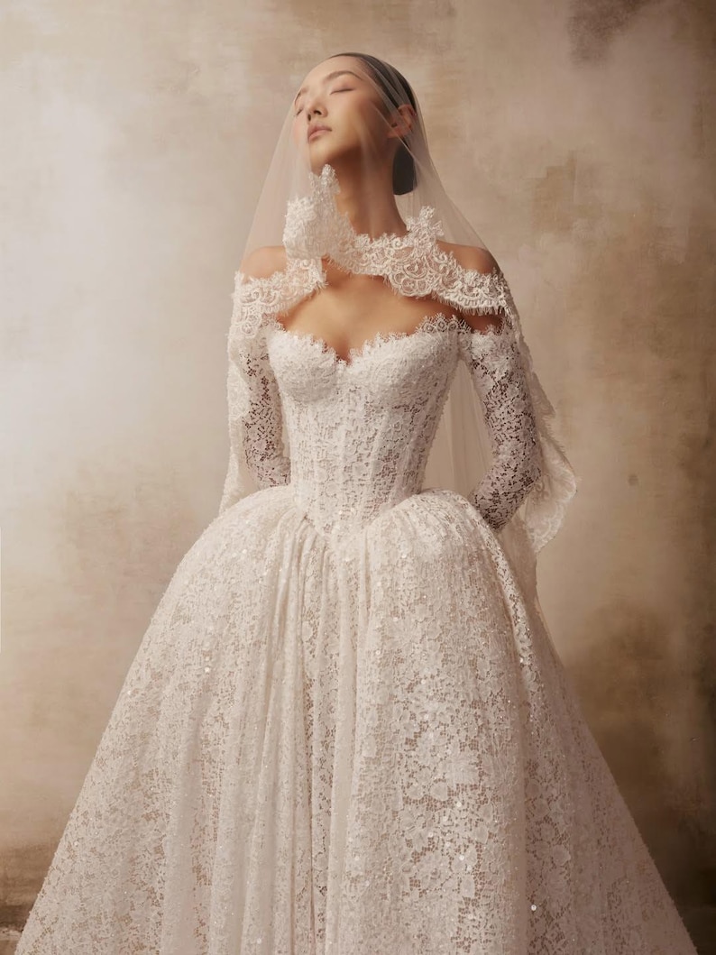 Peut inclure: Une robe en dentelle blanche dot&eacute;e d'un corsage structur&eacute;, de manches longues et d'une encolure d&eacute;gag&eacute;e aux bords festonn&eacute;s. La jupe volumineuse est confectionn&eacute;e en dentelle florale complexe avec de fines paillettes et un voile transparent.
