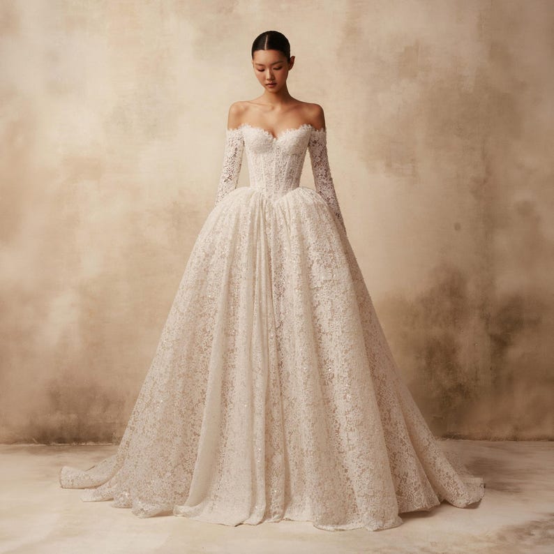 Peut inclure: Une robe longue &agrave; &eacute;paules d&eacute;nud&eacute;es dot&eacute;e d'un corsage style corset et de manches longues. La robe est confectionn&eacute;e en dentelle blanche avec des motifs floraux complexes et de subtiles d&eacute;corations &agrave; paillettes, formant une silhouette volumineuse.