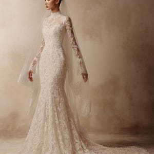 Peut inclure: Une robe de mari&eacute;e en dentelle blanche longue jusqu'au sol avec un col montant, des manches longues et un corsage ajust&eacute; qui s'&eacute;vase en une silhouette sir&egrave;ne avec une tra&icirc;ne. Un voile transparent avec bordure en dentelle est port&eacute;.