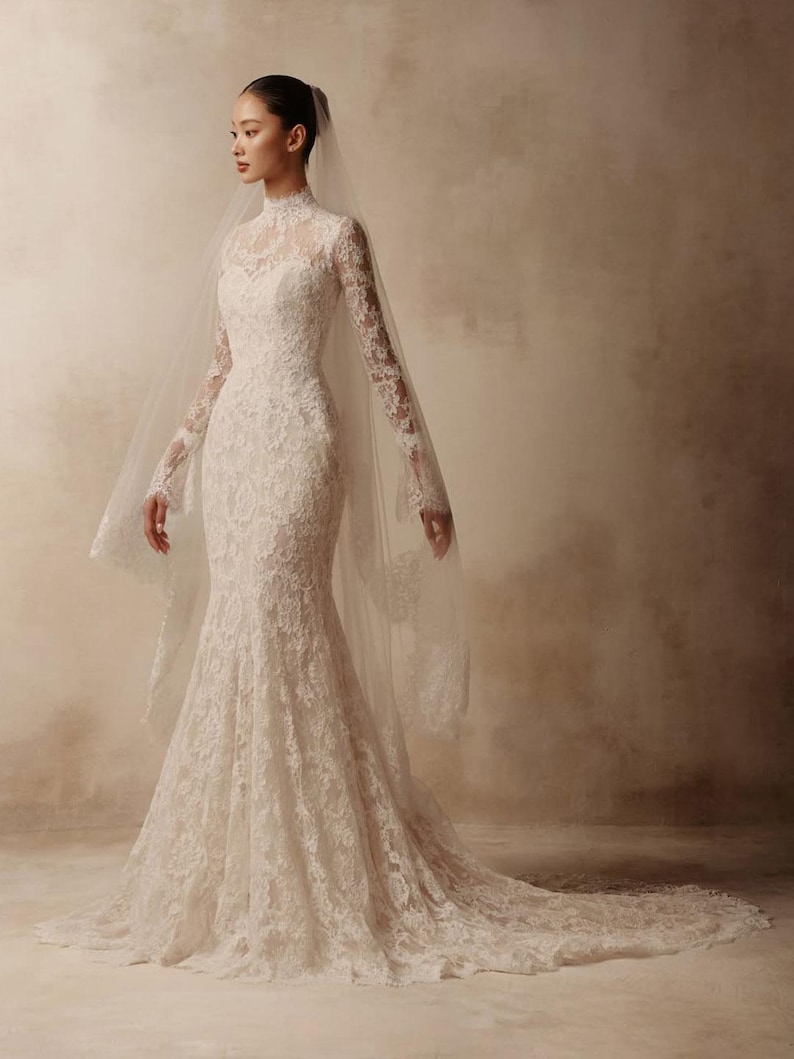 Peut inclure: Une robe de mari&eacute;e en dentelle blanche longue jusqu'au sol avec un col montant, des manches longues et un corsage ajust&eacute; qui s'&eacute;vase en une silhouette sir&egrave;ne avec une tra&icirc;ne. Un voile transparent avec bordure en dentelle est port&eacute;.
