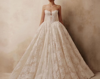 May include: A floor-length white ball gown featuring a strapless corset bodice with a deep center plunge and intricate lace overlay. The voluminous skirt is crafted from layers of fabric with a matching floral lace pattern extending to the hem.