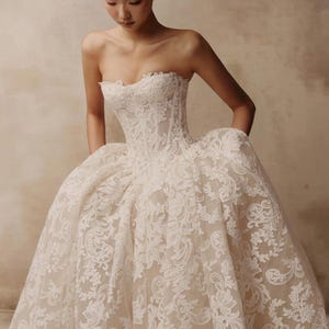 May include: A strapless ball gown featuring a structured corset bodice and a voluminous skirt. The dress is crafted from a nude-toned base fabric overlaid with intricate white floral lace embroidery that extends from the bodice to the hem.