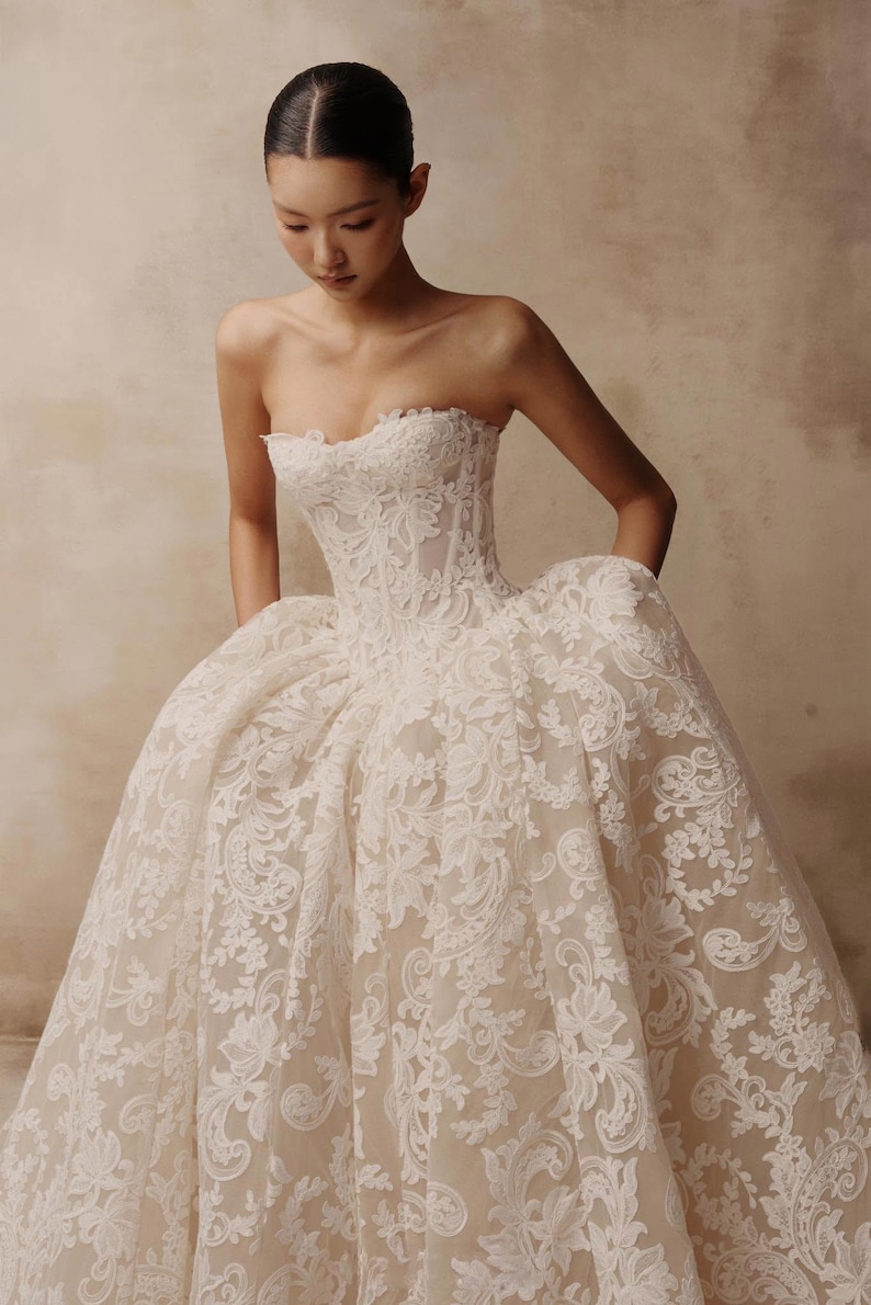 May include: A strapless ball gown featuring a structured corset bodice and a voluminous skirt. The dress is crafted from a nude-toned base fabric overlaid with intricate white floral lace embroidery that extends from the bodice to the hem.