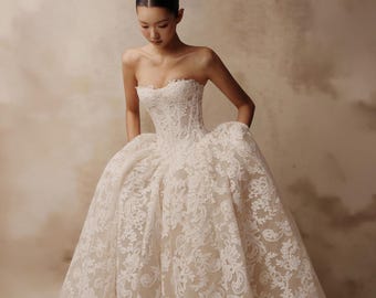 Op de afbeelding: Een strapless baljurk met een nauwsluitend korsetlijfje en een volumineuze rok. De jurk is gemaakt van een transparante, huidkleurige stof die is bedekt met ingewikkeld wit bloemenkantborduurwerk dat doorloopt van het lijfje tot de zoom.