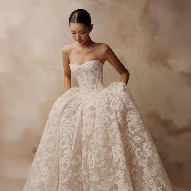 May include: A strapless ball gown featuring a fitted corset bodice and a voluminous skirt. The dress is crafted from a sheer nude-toned fabric overlaid with intricate white floral lace embroidery that extends from the bodice to the hemline.