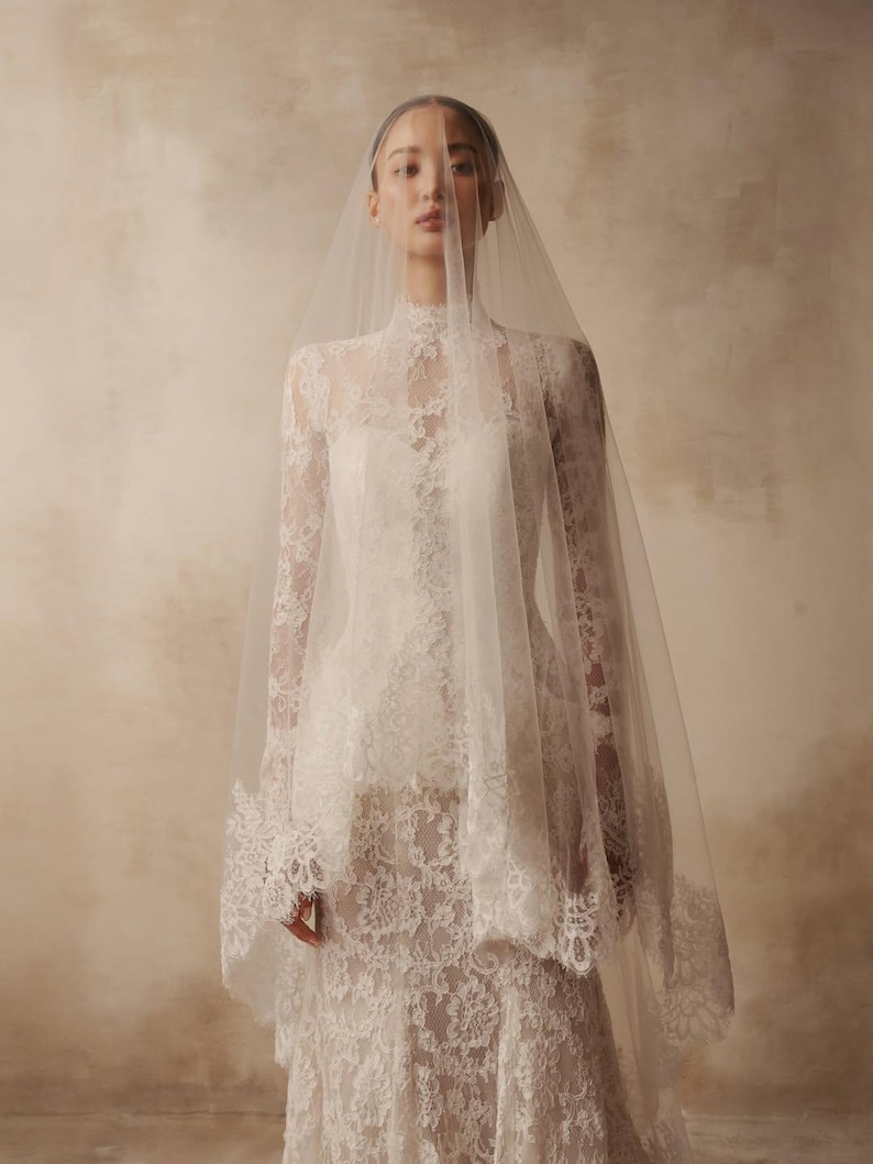 Peut inclure: Une robe longue en dentelle blanche avec un col montant et des manches longues, accompagn&eacute;e d'un voile en tulle l&eacute;ger et transparent qui retombe sur la t&ecirc;te et les &eacute;paules. Un motif floral complexe recouvre le corsage et les manches.