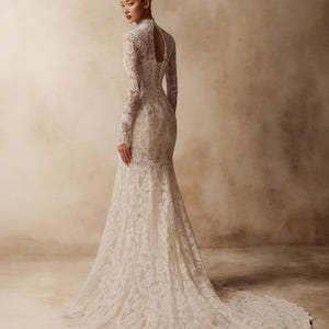 Peut inclure: Une robe longue en dentelle blanche avec des manches longues, un col montant et une ouverture en forme de goutte dans le haut du dos. La robe pr&eacute;sente un corsage ajust&eacute; avec un la&ccedil;age de type corset et une jupe &eacute;vas&eacute;e se terminant par une tra&icirc;ne.