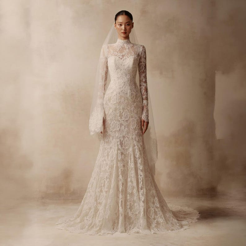 Peut inclure: Une robe longue en dentelle ivoire avec un col montant, des manches longues ajust&eacute;es et une silhouette sir&egrave;ne subtile. La robe est confectionn&eacute;e dans un tissu en dentelle florale complexe et accompagn&eacute;e d'un voile transparent qui tra&icirc;ne.