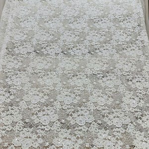 Peut inclure: Un tissu en dentelle blanche pr&eacute;sentant un motif floral complexe sur toute la mati&egrave;re. Le design en maille d&eacute;licate est orn&eacute; de petites paillettes r&eacute;fl&eacute;chissantes dispers&eacute;es sur la surface, ajoutant un &eacute;clat subtil au textile.