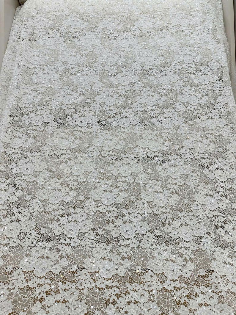 Peut inclure: Un tissu en dentelle blanche pr&eacute;sentant un motif floral complexe sur toute la mati&egrave;re. Le design en maille d&eacute;licate est orn&eacute; de petites paillettes r&eacute;fl&eacute;chissantes dispers&eacute;es sur la surface, ajoutant un &eacute;clat subtil au textile.