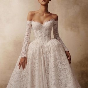 Peut inclure: Une robe blanche longue jusqu'au sol, &agrave; &eacute;paules d&eacute;nud&eacute;es, dot&eacute;e de manches longues en dentelle, d'un corsage structur&eacute; avec un d&eacute;collet&eacute; en c&oelig;ur et d'une jupe volumineuse en dentelle florale complexe agr&eacute;ment&eacute;e de fines paillettes.