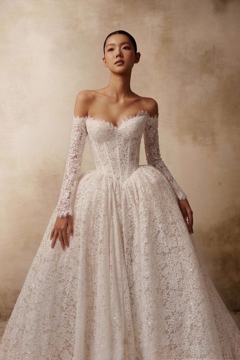 Peut inclure: Une robe blanche longue jusqu'au sol, &agrave; &eacute;paules d&eacute;nud&eacute;es, dot&eacute;e de manches longues en dentelle, d'un corsage structur&eacute; avec un d&eacute;collet&eacute; en c&oelig;ur et d'une jupe volumineuse en dentelle florale complexe agr&eacute;ment&eacute;e de fines paillettes.