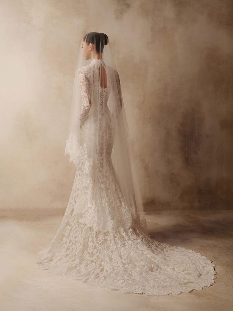 Peut inclure: Une robe de mari&eacute;e en dentelle blanche longue jusqu'au sol avec des manches longues, un col montant et une ouverture en forme de goutte au dos. La robe pr&eacute;sente une superposition de dentelle florale, un corsage ajust&eacute; et une longue tra&icirc;ne.
