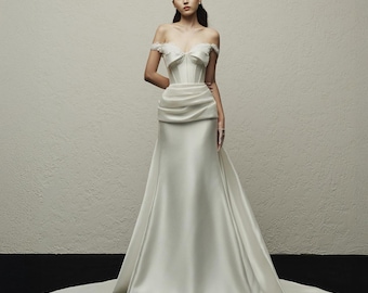 May include: A white satin wedding gown with an off-the-shoulder neckline and a fitted bodice. The dress features a draped waist and a flowing skirt that extends into a train. The model is wearing a headpiece.