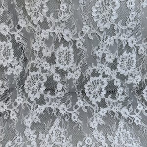 Peut inclure: Un tissu en dentelle blanche pr&eacute;sentant un motif floral complexe avec des bords festonn&eacute;s. Le design se compose de fleurs d&eacute;taill&eacute;es et de vignes entrelac&eacute;es reli&eacute;es par un filet en maille fine semi-transparente, cr&eacute;ant un aspect textur&eacute;.