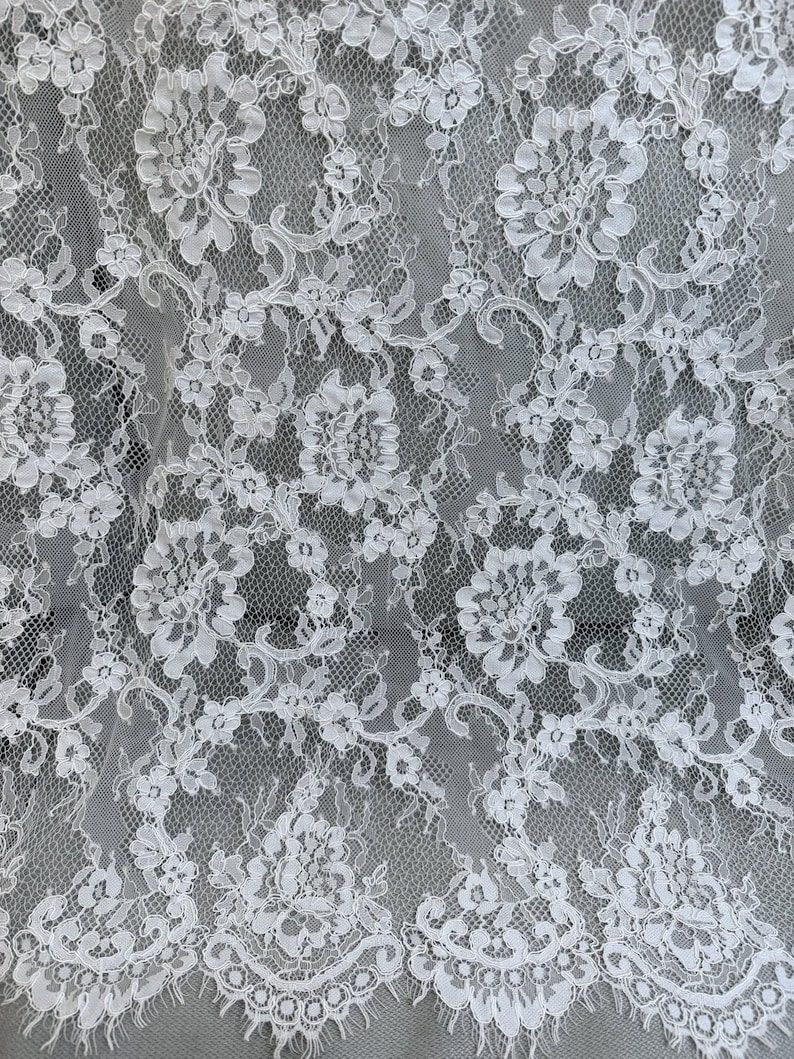Peut inclure: Un tissu en dentelle blanche pr&eacute;sentant un motif floral complexe avec des bords festonn&eacute;s. Le design se compose de fleurs d&eacute;taill&eacute;es et de vignes entrelac&eacute;es reli&eacute;es par un filet en maille fine semi-transparente, cr&eacute;ant un aspect textur&eacute;.