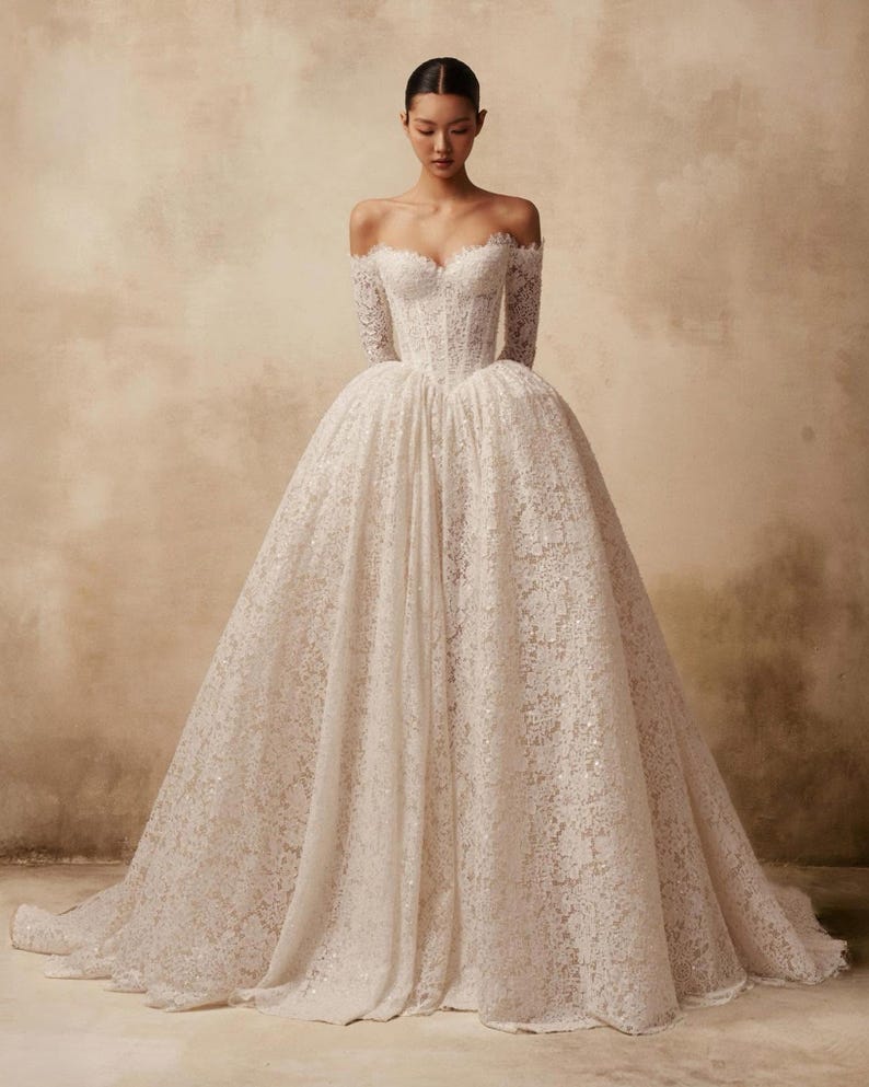 Peut inclure: Une robe longue &agrave; &eacute;paules d&eacute;nud&eacute;es dot&eacute;e d'un corsage ajust&eacute; et d'une jupe volumineuse. L'ensemble du v&ecirc;tement est confectionn&eacute; en dentelle blanche avec des motifs floraux complexes et de subtiles d&eacute;corations &agrave; paillettes sur tout le tissu.