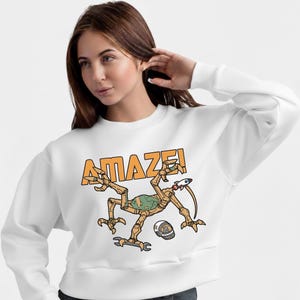 Może przedstawiać: Biała bluza z okrągłym dekoltem z napisem "AMAZE!" w pomarańczowych literach. Grafika przedstawia kresk&oacute;wkowego kosmitę z rakietą, kluczem i hełmem. Kosmita jest brązowy, zielony i beżowy.
