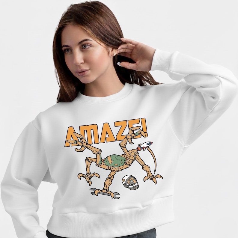 Może przedstawiać: Biała bluza z okrągłym dekoltem z napisem "AMAZE!" w pomarańczowych literach. Grafika przedstawia kresk&oacute;wkowego kosmitę z rakietą, kluczem i hełmem. Kosmita jest brązowy, zielony i beżowy.