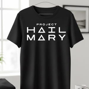 Może przedstawiać: Czarny t-shirt z białym napisem "PROJECT HAIL MARY" pogrubioną czcionką bezszeryfową. Koszulka wisi na metalowym wieszaku. Projekt jest wyśrodkowany na klatce piersiowej.