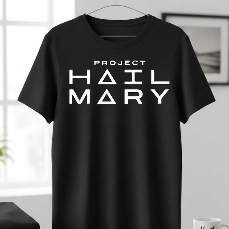 Może przedstawiać: Czarny t-shirt z białym napisem "PROJECT HAIL MARY" pogrubioną czcionką bezszeryfową. Koszulka wisi na metalowym wieszaku. Projekt jest wyśrodkowany na klatce piersiowej.