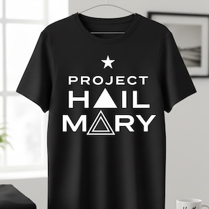 Może przedstawiać: Czarny t-shirt z białym napisem "PROJECT HAIL MARY" i gwiazdą nad nim. Koszulka wisi na metalowym wieszaku. Projekt zawiera tr&oacute;jkąt i kwadrat.