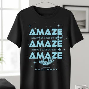 Może przedstawiać: Czarna koszulka z napisem "AMAZE" w jasnoniebieskim kolorze, powt&oacute;rzonym trzy razy z grafiką o tematyce kosmicznej. Tekst "PROJECT HAIL MARY" znajduje się pod grafiką szkieletu dłoni.