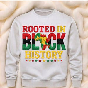 Puede incluir: Sudadera blanca con el texto "ROOTED IN BLACK HISTORY" en rojo, negro, verde y amarillo. El diseño incluye un mapa de África. El texto está bordado con un diseño de estrellas en la parte inferior.