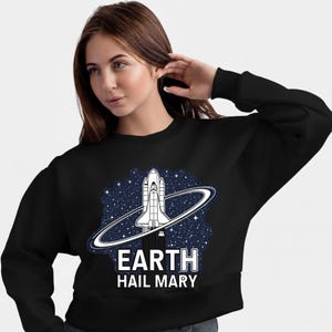 Może przedstawiać: Czarna bluza z białym nadrukiem przedstawiającym prom kosmiczny z pierścieniem na tle gwiezdnego nieba. Pod spodem widnieje napis "EARTH HAIL MARY". Bluza ma okrągły dekolt.