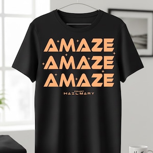 Może przedstawiać: Czarny t-shirt z pomarańczowym napisem "AMAZE" powt&oacute;rzonym trzy razy, z gwiazdkami. Tekst "PROJECT HAIL MARY" jest wydrukowany poniżej. Koszulka wisi na metalowym wieszaku.