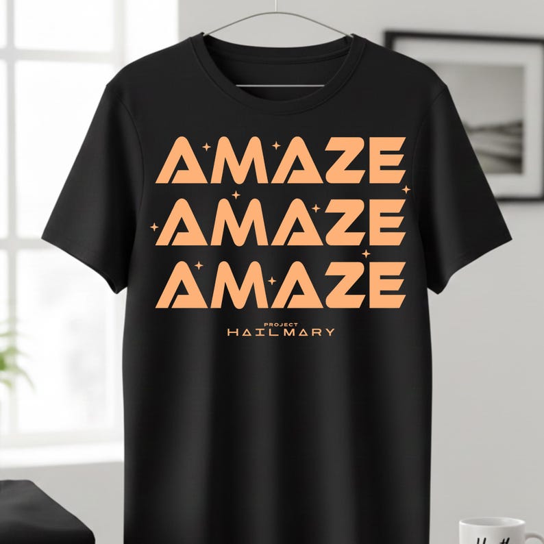 Może przedstawiać: Czarny t-shirt z pomarańczowym napisem "AMAZE" powt&oacute;rzonym trzy razy, z gwiazdkami. Tekst "PROJECT HAIL MARY" jest wydrukowany poniżej. Koszulka wisi na metalowym wieszaku.