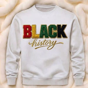 May include: White sweatshirt with the words "BLACK history" in colorful, textured lettering. The word "BLACK" is in red, yellow, green, and black. The word "history" is in gold script.