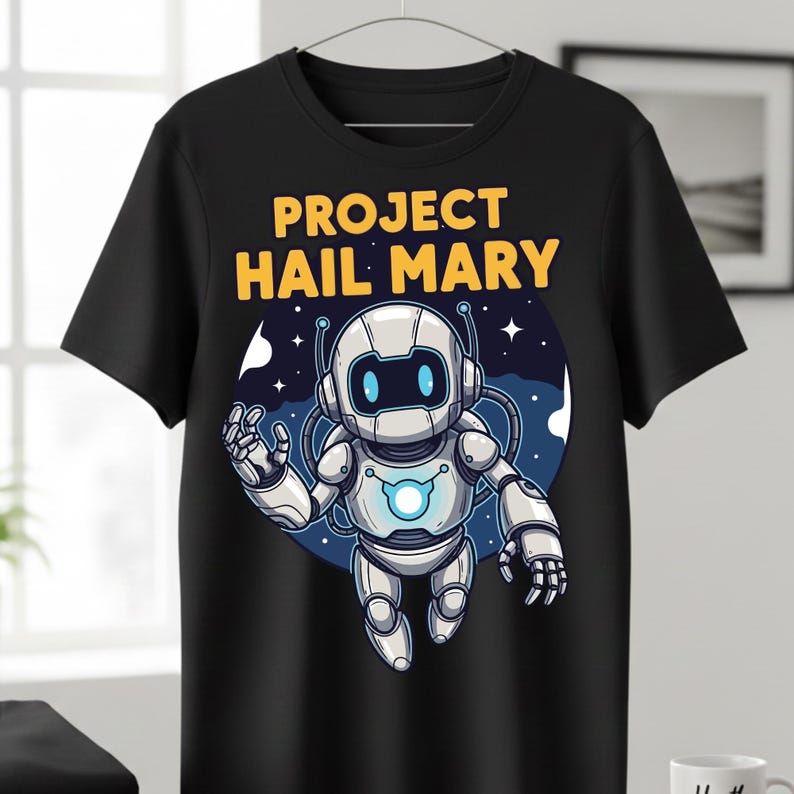 Może przedstawiać: Czarny t-shirt z kresk&oacute;wkowym robotem i ż&oacute;łtym napisem "PROJECT HAIL MARY". Robot jest biało-srebrny z niebieskimi akcentami, na niebiesko-białym tle z gwiazdami. Projekt jest umieszczony na środku koszulki.