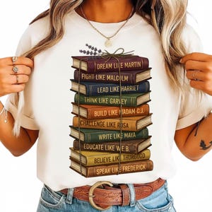 Puede incluir: Camiseta blanca con una pila de libros coloridos, cada uno con una cita inspiradora y un nombre como Martin o Malcolm. Los libros están atados con hilo y una ramita de lavanda.