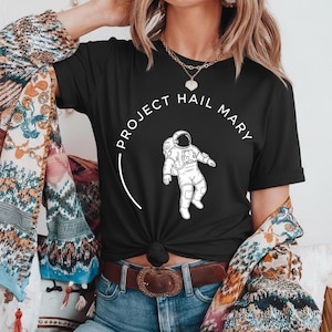 Może przedstawiać: Czarny t-shirt z białą grafiką astronauty i napisem "PROJECT HAIL MARY" w okrągłym wzorze. Koszulka ma kr&oacute;tkie rękawy i jest wykonana z miękkiego materiału. Projekt znajduje się z przodu.