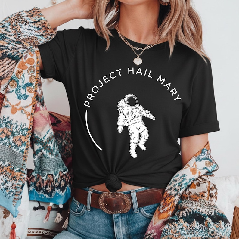 Może przedstawiać: Czarny t-shirt z białą grafiką astronauty i napisem "PROJECT HAIL MARY" w okrągłym wzorze. Koszulka ma kr&oacute;tkie rękawy i jest wykonana z miękkiego materiału. Projekt znajduje się z przodu.