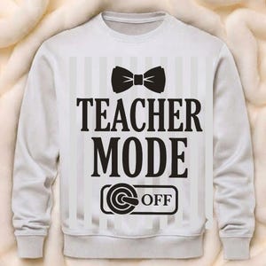May include: White sweatshirt featuring a black bow tie graphic and the words "TEACHER MODE OFF" in black. The design includes a striped background. A casual, comfortable garment for teachers.