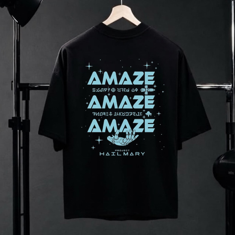 Może przedstawiać: Czarny t-shirt ze słowem "AMAZE" w jasnoniebieskim kolorze, powt&oacute;rzonym trzy razy r&oacute;żnymi czcionkami. Projekt zawiera grafikę dłoni i tekst "PROJECT HAILMARY".