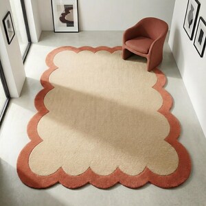 May include: A large, beige rug with a scalloped, coral-colored border. A matching coral-colored armchair sits on the rug. The rug is in a room with white walls and framed artwork.