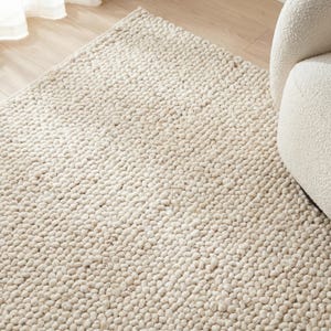 Puede incluir: Alfombra de área texturizada de color crema con un pelo denso y en bucle. La alfombra tiene un tono suave y neutro y una apariencia táctil. La alfombra está colocada sobre un suelo de madera clara, con parte de una silla blanca visible.