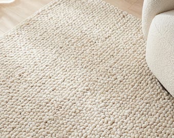 May include: Cream-colored, textured area rug with a dense, looped pile. The rug has a soft, neutral tone and a tactile appearance. The rug is placed on a light-colored wooden floor, with a portion of a white chair visible.