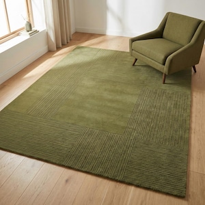 May include: A large, olive green area rug with a geometric design. The rug features a central solid section surrounded by patterned borders with linear textures. A matching green armchair sits on the rug.
