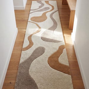 May include: A rectangular hallway rug with an abstract design. The rug features a cream background with flowing, organic shapes in shades of brown and grey. The rug is placed on a wooden floor, and the hallway is flanked by white walls.