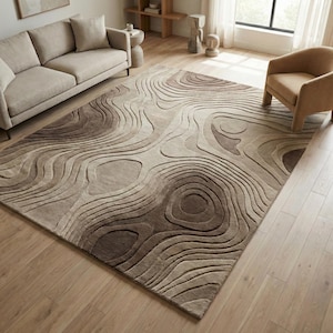 May include: A large, rectangular area rug with a topographic design in shades of brown and beige. The rug features a raised, layered pattern resembling contour lines, creating a textured surface. The rug is placed on a hardwood floor in a living room setting.
